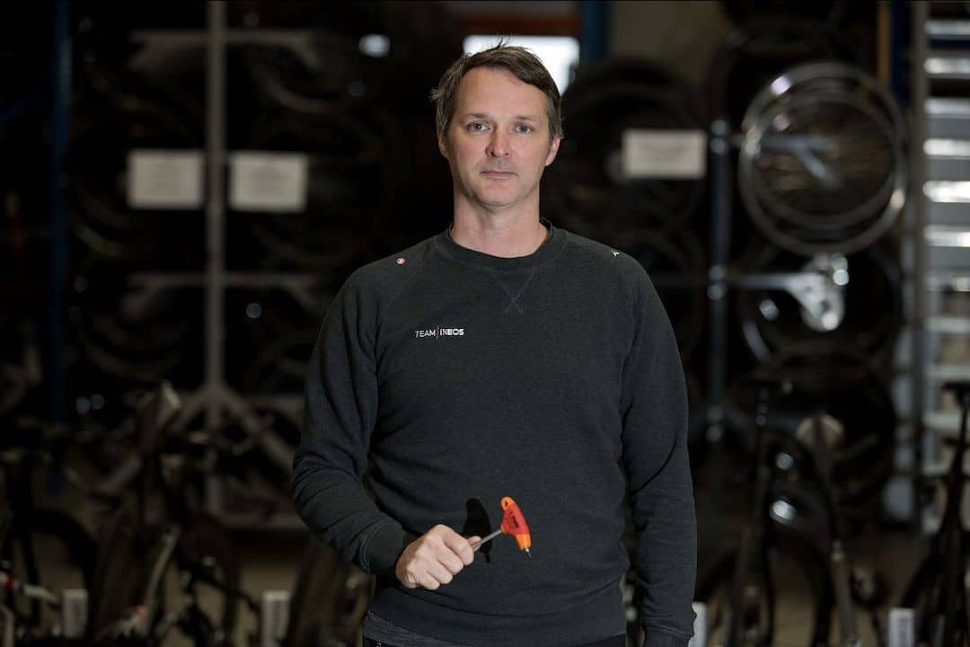 Team Ineos mechanic in service-course apron holding a red and orange Unior screwdriver, team bikes racked behind him