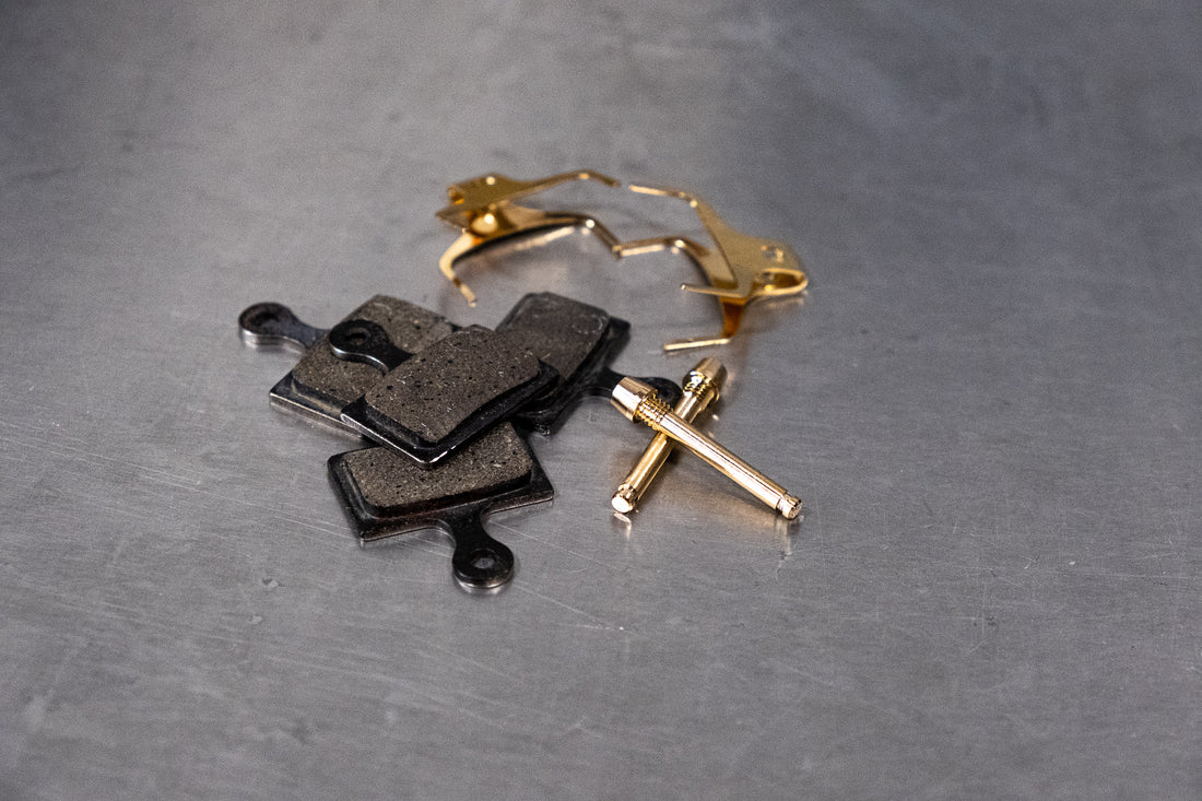 Worn black disc brake pads and a gold caliper retaining pin set on a metal workshop bench before bedding-in