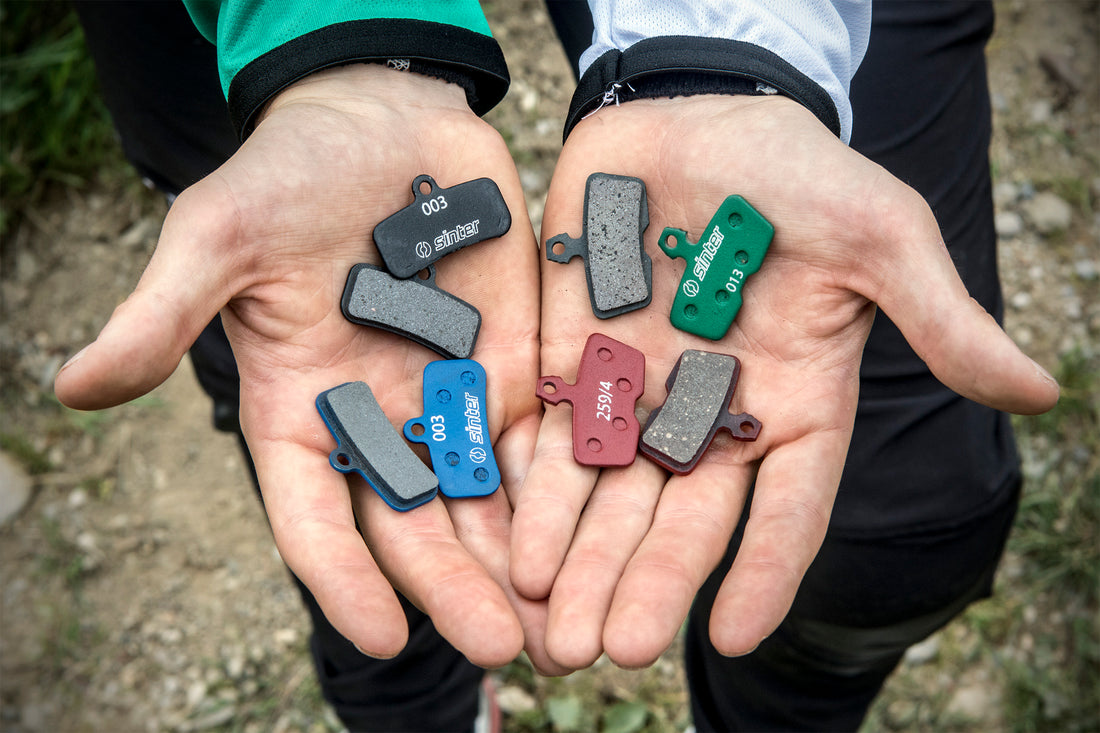 Eight Sinter disc brake pads in two cupped hands, showing black, green, blue, and red compounds with stamped s-codes