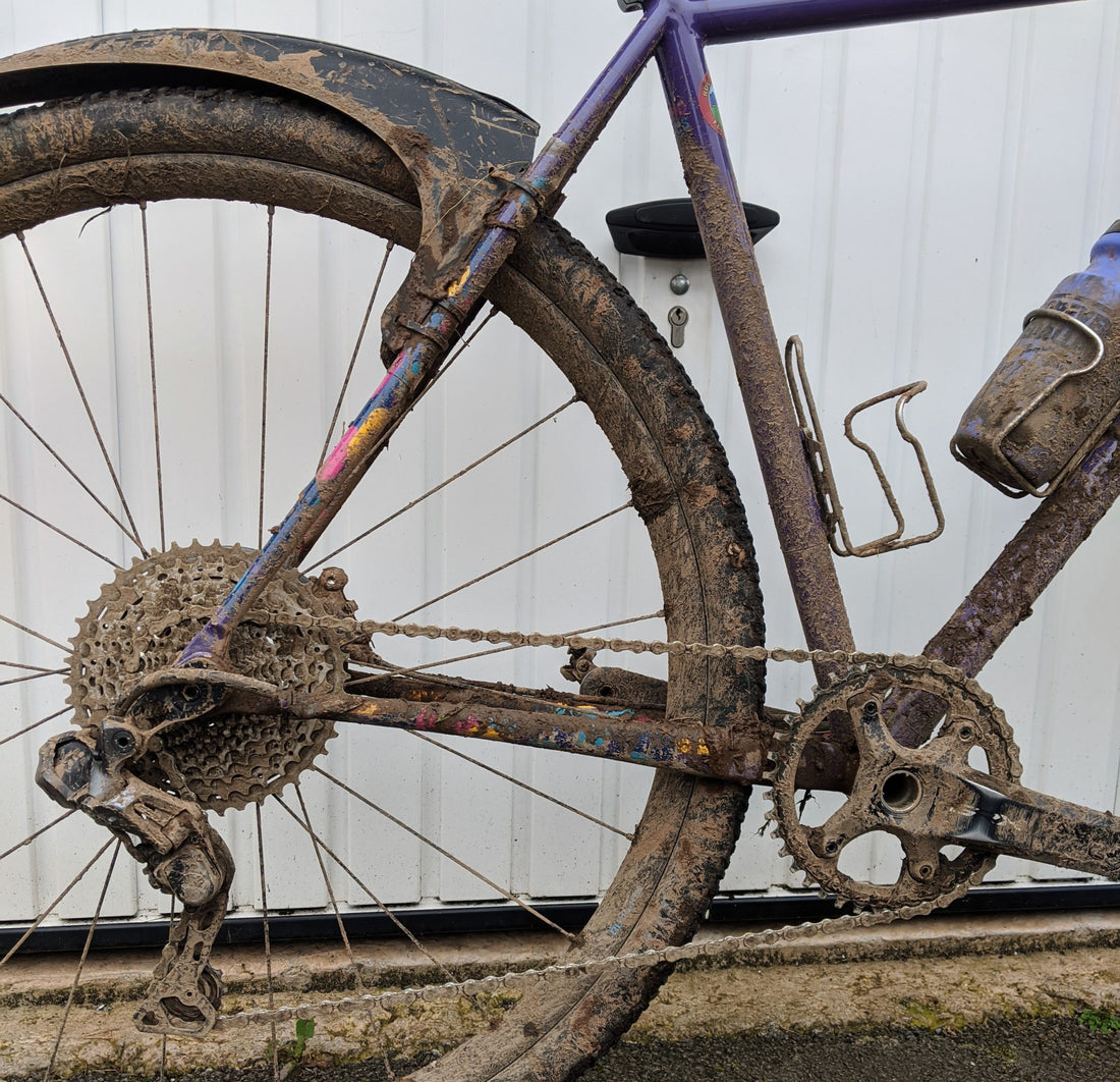 Mud-caked cassette, chain, and chainrings on a gravel bicycle after a wet ride, leaning against a corrugated wall
