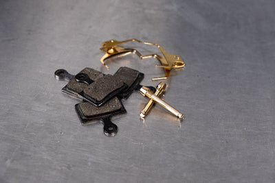 Worn black disc brake pads and a gold caliper retaining pin set on a metal workshop bench before bedding-in