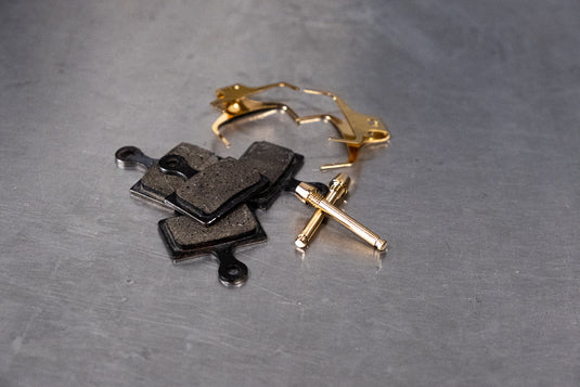 Worn black disc brake pads and a gold caliper retaining pin set on a metal workshop bench before bedding-in