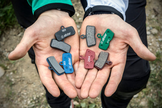 Eight Sinter disc brake pads in two cupped hands, showing black, green, blue, and red compounds with stamped s-codes