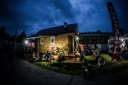 Grinduro 2019 race village at dusk, Devinci flag and cyclists relaxing outside a wooden cabin with parked bicycles
