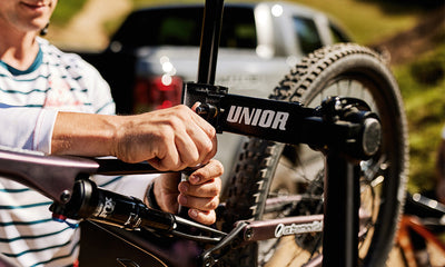 Mechanic clamping a mountain bike into a Unior repair stand beside a pickup truck, hand tightening the clamp jaw