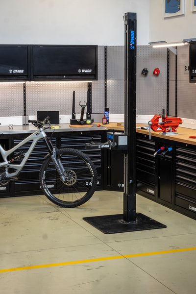 Unior Electric Repair Stand 1693EL in a workshop with a bicycle clamped, Unior cabinets and pegboards behind — representative e-bike service-bay setup.