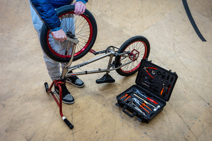 Rider flipping a BMX bicycle on a folding stand at a skatepark, open Unior BMX Tool Kit 1600BMX case nearby