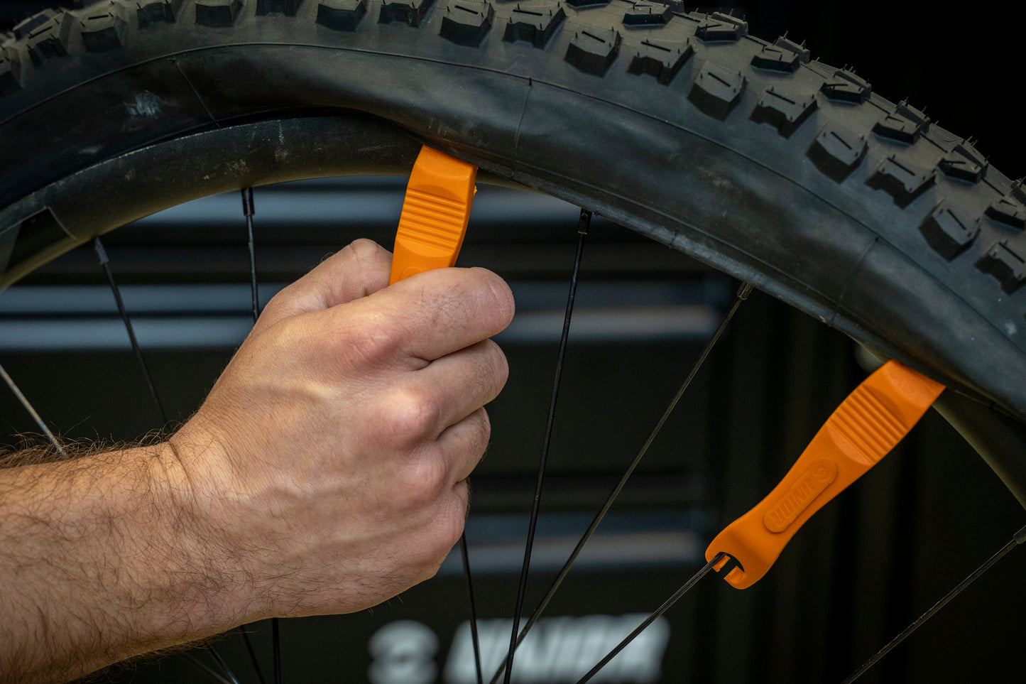 Orange Unior 1657 tire lever hooked onto a spoke holding the bead off the rim while a second lever walks the tire free