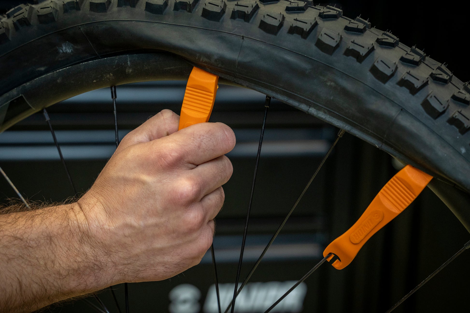 Orange Unior 1657 tire lever hooked onto a spoke holding the bead off the rim while a second lever walks the tire free