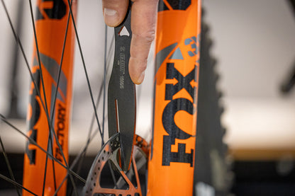 Rotor Truing Fork 2.0 held next to an orange Fox Factory mountain bike fork and disc rotor for an alignment check