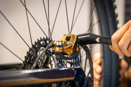 Rotor Truing Fork 2.0 engaged with a mountain bike disc rotor at the gold brake caliper, cassette and wheel in frame