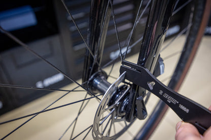 Rotor Truing Fork 2.0 hooked around a road bicycle disc rotor at the front hub, workshop drawer cabinet behind