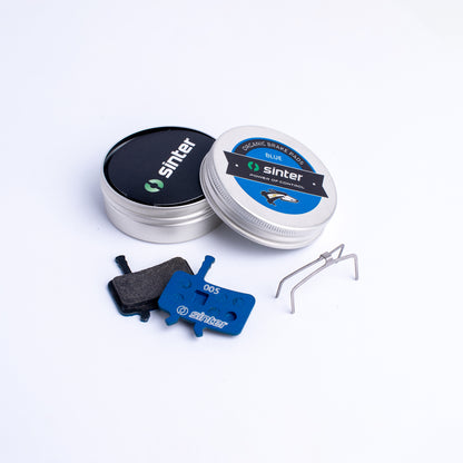 Sinter Model 005 Blue organic disc brake pads with retention spring beside aluminium tin packaging for Avid calipers