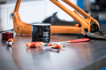 Black Unior enameled mug printed Made for Work on a workshop bench beside Unior screwdrivers and an orange mountain bike frame