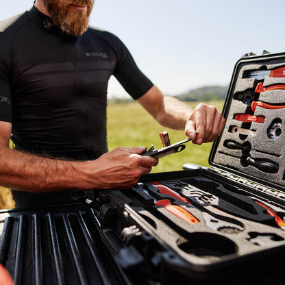 Cyclist in black jersey using a tool from the open Unior Home Kit case on a truck tailgate in an open field